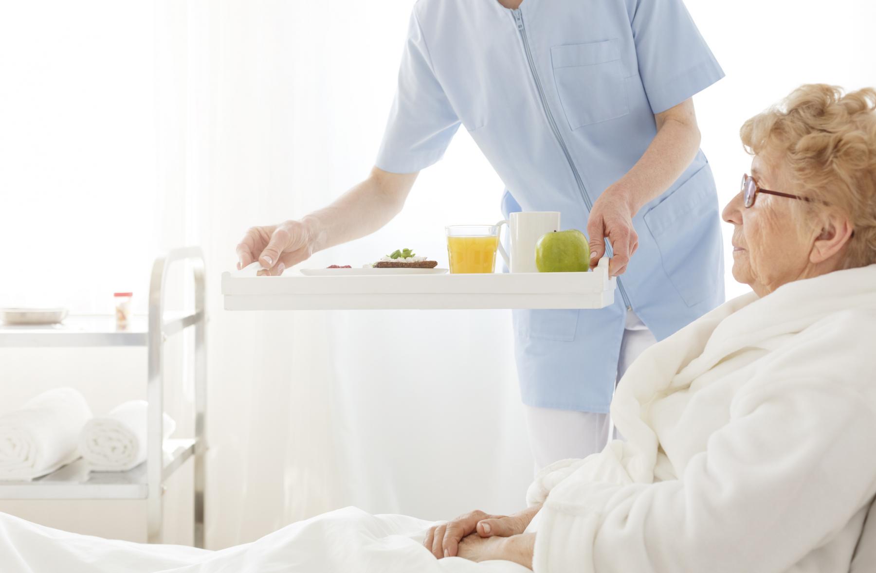 A patient lying in a hospital bed is served a tray of food
